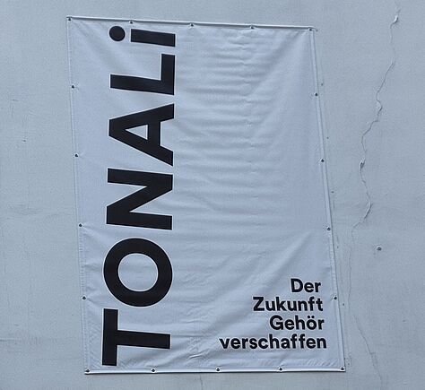 Ein weißes Banner ist an einer weißen Mauer befestigt (draußen). Links senkrecht von unten nach oben: TOMALi. Rechts unten in der Ecke: Der Zukunft Gehör Verschaffen. Jedes Wort in einer neuen Zeile.