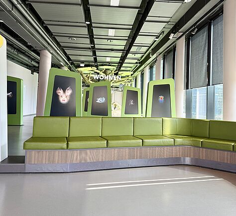 Im Vordergrund steht eine geschwungene Sitzbank mit grünen Polstern. Hinter der Sitzbank sind mehrere große, grün gerahmte Tafeln mit Tiermotiven aufgestellt, links daneben führt eine Stufe zu dem Raum dahinter. Über den Tafeln hängt das Wort 'WOHNEN' in weißen Buchstaben.