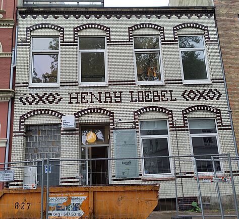Weiße Fassade eines Altbau-Wohnhauses mit Ziegelverzierungen in dunkelbraun. 6 hohe Fenster mit Rundbögen, auf 2 Stockwerke verteilt. Schriftzug „HENRY LOEBEL“ mittig über den Fenstern im ersten Stock. Das zweite von links der 3 unteren ist die Eingangstür. Das links daneben ist mit Glasbausteinen gefüllt. Vor dem Gebäude ein Bauzaun und ein Schuttcontainer.