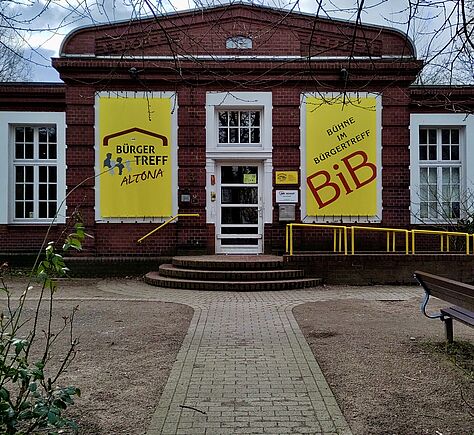 Der Eingangsbereich des einstöckigen Gebäudes. Links und rechts von der Tür zwei große gelbe Plakate. Auf den Plakaten steht Bürgertreff und BIB. Zur Eingangstür führen drei Stufen. Rechts eine Rampe mit einem gelben Handlauf. Im Vordergrund der gepflasterte Gehweg.