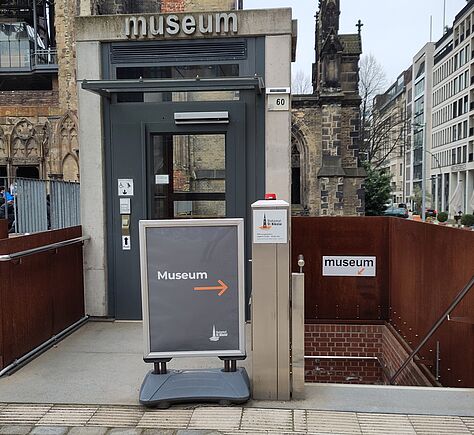 Ein graues Schild auf einem Ständer mit der Aufschrift „Museum“ und einem orangefarbenen Pfeil nach rechts. Darüber steht „Museum“ in hellen Buchstaben über einer grauen Tür. Rechts ist ein weiteres kleines Schild mit Pfeil, das zu einer Treppe nach unten führt.