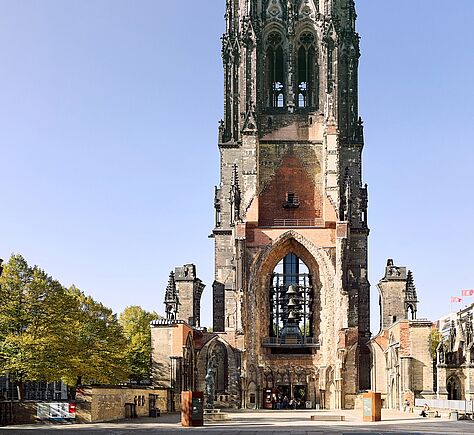 Ein hoher, teilweise zerstörter Kirchturm aus hellem und dunklem Stein steht auf einem offenen Platz. Die Fassade wirkt rau und alt, mit vielen Details und großen Öffnungen. Der Platz davor ist weit und ruhig, mit Bäumen an der Seite und einer klaren, hellen Stimmung.
