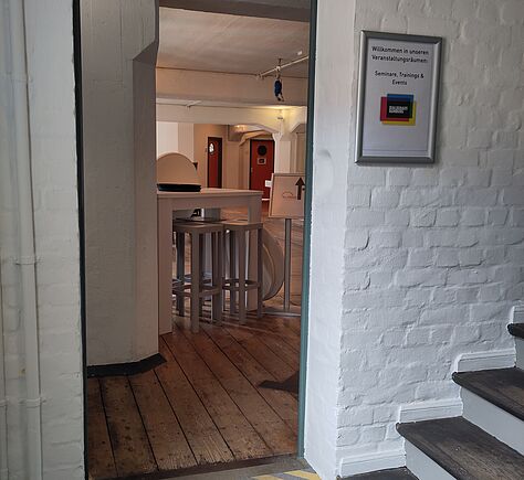 Die Eingangstür zum Café hat eine gelbschwarz gestreifte Stufe. Im Hintergrund auf einem Holzboden stehen im Café hohe weiße Tische und Hocker. Im Vordergrund rechts einige Treppenstufen.
