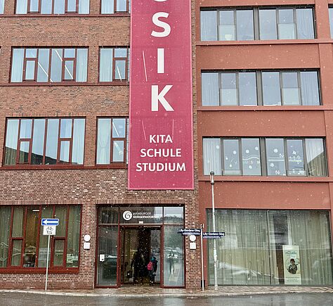 Ein mehrstöckiges Gebäude aus rotem Backstein mit vielen großen Fenstern. In der Mitte hängt ein langes rotes Banner mit der Aufschrift „Musik“ (vertikal geschrieben) sowie „Kita, Schule, Studium" (horizontal). Unten befindet sich der Eingang zum Hamburger Konservatorium.