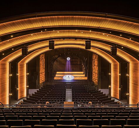 Der leere Theatersaal in einer Totalen. In der Mitte der Bühne eine große Uhr, die sich spiegelt. Golden scheinen Lichtbögen in Form der Bühne im ganzen Saal.
