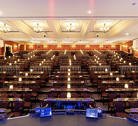 Das Foto ist von der Bühne aus in einen festlich beleuchteten, nach hintern aufsteigenden Theatersaal mit vielen Reihen schmaler Tische und Stühle aufgenommen. Auf jedem Tisch steht eine kleine Lampe mit warmem Licht. Die Wände sind mit roten Tapeten und goldenen Verzierungen gestaltet. An der Decke hängen mehrere kleine Kronleuchter. Im Vordergrund befinden sich technische Geräte auf der Bühnenkante. In Hintergrund links oben ist eine Tür.