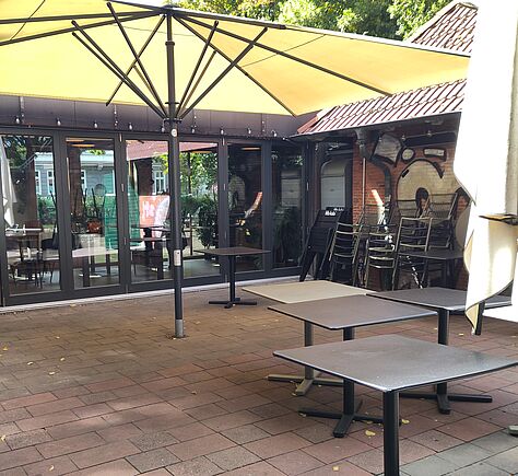 Auf einer Terrasse mit rötlichen und grauen Pflastersteinen stehen mehrere rechteckige Tische. Ein großer, gelber, quadratischer Sonnenschirm überspannt den Bereich. Im Hintergrund sind bodentiefe Fenster und eine Tür, rechts stehen gestapelte Stühle. Hinter dem Gebäude sind hohe belaubte Bäume.