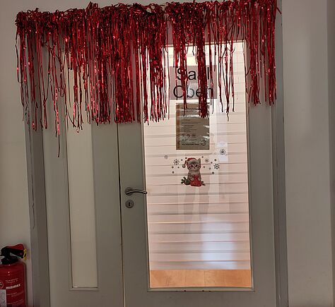 Eine weiße Zimmer-Flügeltür mit Glasscheibe, der Rahmen ist im oberen Teil mit roten Fransen dekoriert. Links im Türrahmen ein schmaler Flügel ohne Klinke mit Glasscheibe.