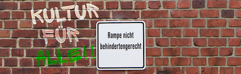 Eine Hauswand mit roten Ziegeln. Darauf ein Schild mit dem Text: Rampe nicht behindertengerecht. Rechts davon gesprüht: Kultur für Alle