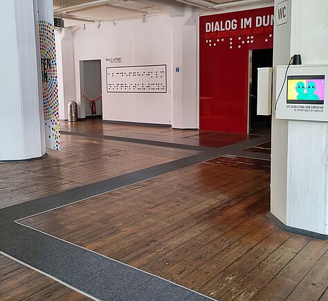 In einem größeren Raum sind breite Leitstreifen tast- und sichtbar auf dem Holzboden. Rechts ist eine rot gerahmte Tür, links daneben auf der weißen Wand ein Text in Braille-Schrift, links daneben eine weitere Tür.
