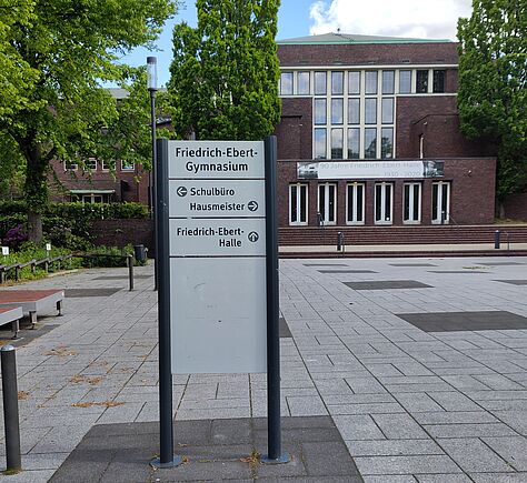 Ein großer gepflasterter Vorplatz mit großen Gehwegplatten belegt. Symetrisch sind Rechtecke dunkel abgesetzt. Im Vordergrund auf einem dunklen Rechteck eine weiße Hinweistafel zwischen 2 Ständern, die mit schwarzen Pfeilen auf auf Schulbüro, Hausmeister und Friedrich-Ebert-Halle hinweist. Im Hintergrund das große Backsteingebäuder der FEH, breite Stufen davor. An den Seiten hohe Bäume mit frischem Laub.