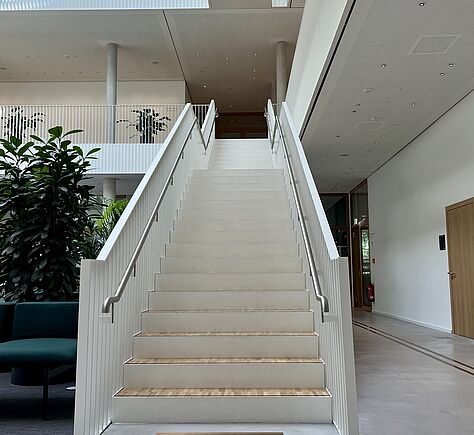 Die helle Treppe hat vor der ersten Stufe ein Aufmerksamkeits Feld. Jede Stufe ist markiert, links und rechts ein Handlauf. Die Stufen selbst sind aus Holz, die Schutzgitter links und rechts sind weiß gestrichen.