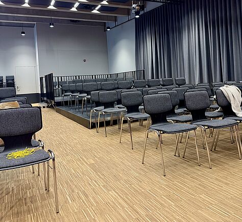 Ein heller Veranstaltungsraum mit Holzboden und vielen grauen Stühlen in Reihen. Ein Teil der Sitzplätze steht auf einer kleinen erhöhten Plattform. An der Seite hängen lange graue Vorhänge und an der Decke sind Lampen und Technik befestigt.
