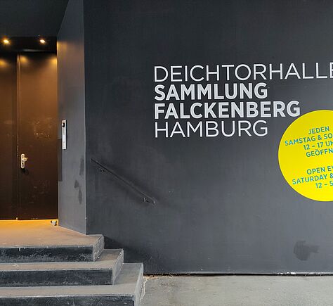 Links führen drei Treppenstufen zu einer schwarzen Tür. Rechts eine schwarze Wand, an der ein Handlauf über der Treppe befestigt ist. Auf der Wand steht in weißer Schrift untereinander 'Deichtorhallen Sammlung Falckenberg Hamburg'. Darunter ein gelber Kreis auf dem in blauer Schrift die Öffnungszeiten stehen.