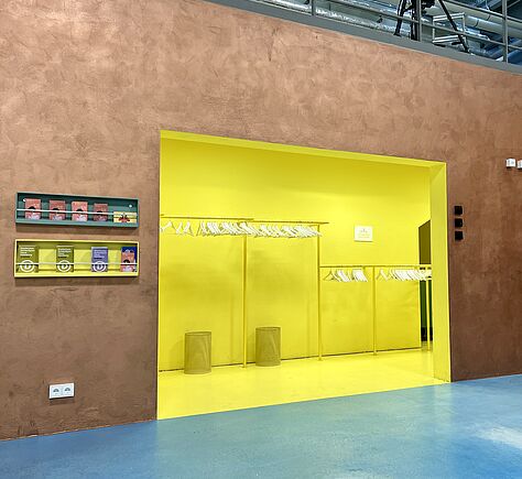 Das Bild zeigt im Foyer die Garderobe. Boden und Wand sind in einem leuchtenden Gelb gestrichen. Links daneben an einer Gold gehaltenen Wand ein schmales Regal mit kleinen Boxen mit Broschüren. In der Garderobe Metallständer in zwei unterschiedlichen Höhen mit vielen weißen Kleiderbügeln.