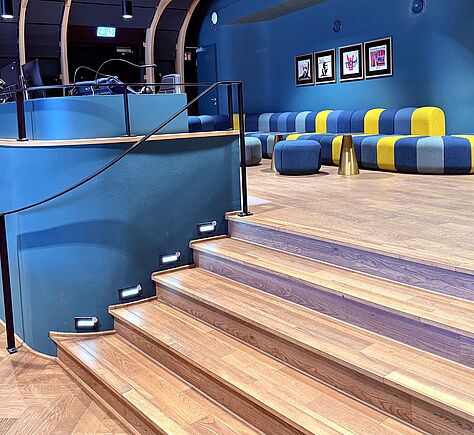 Im Mittelpunkt des Bildes, die Stufen aus Holz, die in das Parkett führen. Oben ist eine Reihe von gepolsterten Sitzbänken und hocken in den Farben blau, hellblau und gelb zu sehen. An der blauen Wand über den Sitzgelegenheiten hängen vier gerahmte Bilder.