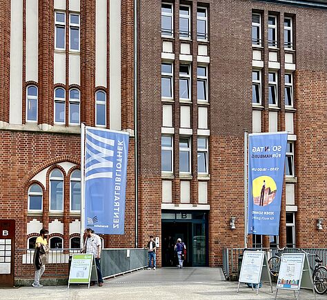 Ein rotes Backsteingebäude mit einem breiten Eingang. Links und rechts davon große Fahnen in hellblau mit dem Logo der Bücherhallen Hamburg.