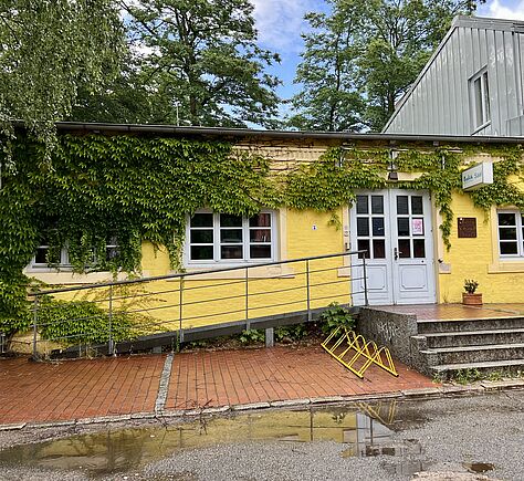 Ein gelb gestrichener einstöckiger Anbau mit einer weißen Tür, der Saaleingang. Links davon eine Rampe mit Geländer. Davor rot gepflasterter Weg, uneben. Vor dem Eingang vier Stufen. Im Vordergrund eine belaubte Baumkrone 