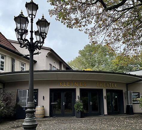 Das Halbrund der Front eines niedrigen Gebäudes mit Flachdach, dessen Rand weit vorspringt. 3 dunkle Eingangstüren sind nebeneinander angeordnet, darüber in goldener großer Schrift: Kleines Theater. Vor dem Gebäude ein gepflasterter Platz, links im Vordergrund eine alte Laterne mit geschmiedetem Pfahl..