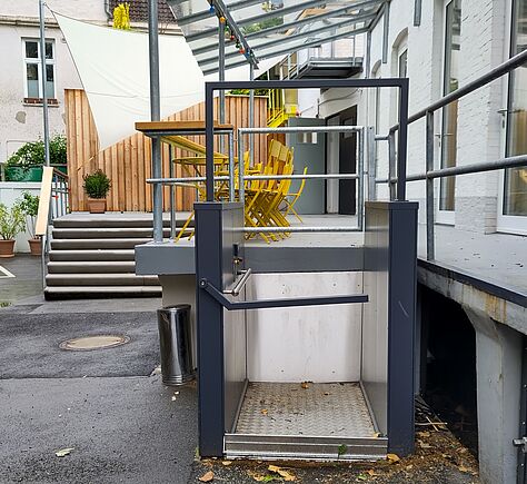 Vor dem Podest steht ein Hublifter. Links davon ist die Treppe zu sehen, im Hintergrund eine Holzwand mit schräger Oberkante. Auf dem Podest stehen gelbe Metalltische und -stühle, darüber eine transparente Überdachungskonstruktion