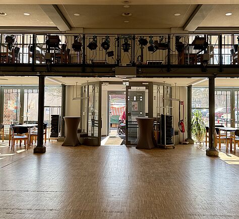 Ein großer, heller Raum mit Holzboden und einem Eingang mit Glastür in der Mitte. Vor der Tür stehen zwei hohe Stehtische, daneben Wagen mit gestapelten Stühlen. Über dem Eingang befindet sich eine Empore mit vielen Scheinwerfern und Technik.