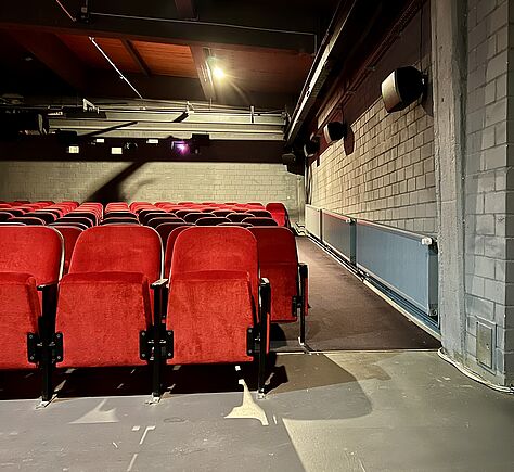 Der Kinosaal mit mehreren Reihen von roten Sitzen, die nach vorne hin Absteigend angeordnet sind. Die Sitze sind leer, an der rechten Seite ist eine Wand mit grauen Ziegeln und darauf montierten Lautsprechern zu sehen. 
