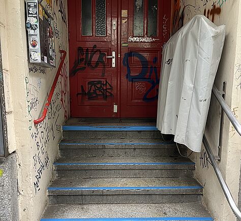 Eine steinerne schmale Treppe mit 6 Stufen führt auf eine rote Eingangstür zu. Rechts ist die Plane, die über einem Treppenlifter hängt zu sehen. Die Stufen sind an der Vorder- und Oberkante mit blauem Klebestreifen markiert.