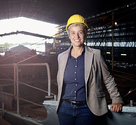 Ursula Richenberger steht in einer Halle auf einer Werft, trägt einen gelben Helm und lacht.