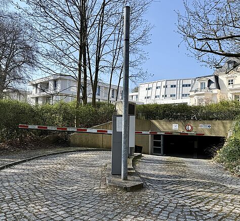 Eine mit Kopfsteinpflaster versehene Zufahrt zu der Tiefgarage, die bergab führt. In der Mitte eine Schranke, die geöffnet senkrecht steht. Links und rechts der Zufahrt sind Grünflächen mit niedrigen Büschen. Im Hintergrund mehrere Gebäude, darunter ein historisch gebautes Haus mit Giebeldach. Der Himmel ist blau.