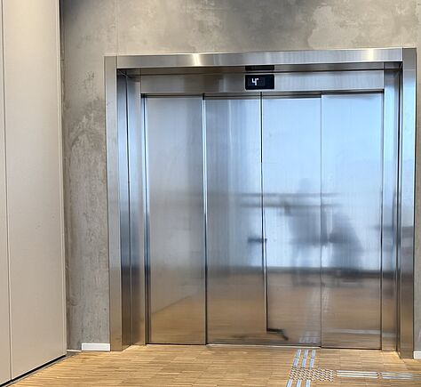 Ein moderner Aufzug mit großen silbernen Metalltüren in einer Betonwand. Über der Tür zeigt eine digitale Anzeige die Zahl „4“. Der Boden davor ist aus Holz und hat tastbare Leitstreifen zur Orientierung.