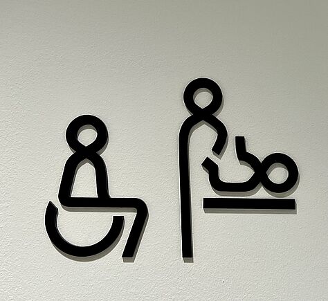 Zwei schwarze Piktogramme auf weißem Untergrund. Rechts eine Person, die ein Baby wickelt. Links ein Rollstuhlsymbol