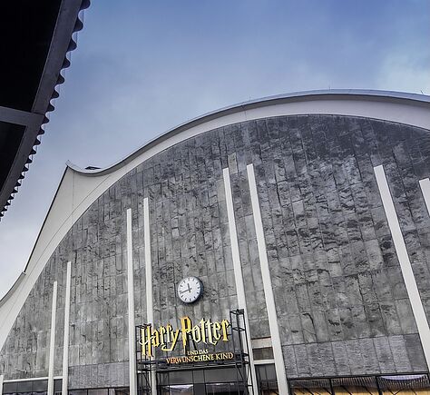 Außenansicht bei Tageslicht. Über dem Eingang der oval geschwungenen Fassade eine große Uhr und der Text: Harry Potter und das verwunschene Kind