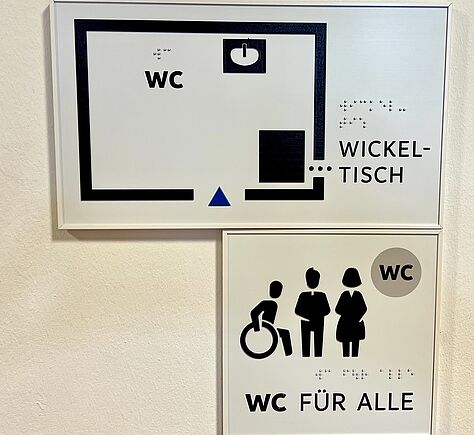 Ein Schild in der Nahaufnahme. WC für alle, mit den Piktogrammen für Rollstuhlfahrer, wann und Frau. Alles ist auch mit Breil beschriftet. Die Objekte im Raum sind in ihrer Position auf dem Schild zu tasten.