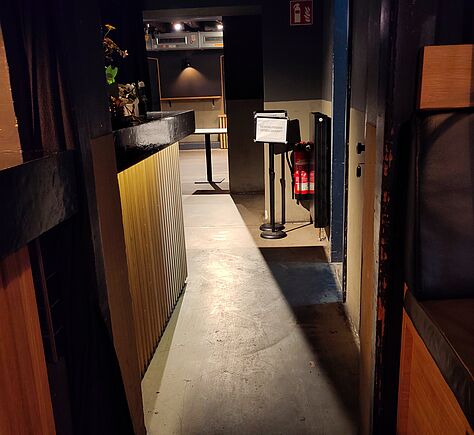 Ein schmaler Gang mit Betonboden führt links zu einem dunklen Tresen mit vertikalen Holzleisten, darauf ein paar Blumen. Rechts steht ein Ständer mit einem Blatt Papier, daneben hängt ein roter Feuerlöscher. Die Wände sind dunkel, im hinteren Raum steht ein Tisch. Die Beleuchtung ist gedämpft, einige Deckenstrahler werfen Licht auf den Boden. 