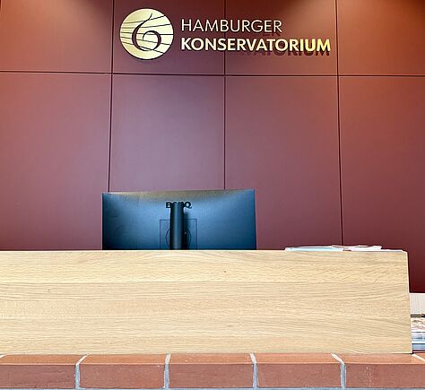 Ein Empfangstresen aus hellem Holz mit einer breiten, glatten Oberfläche. Die Vorderseite besteht aus großen Holzplatten und steht auf einer niedrigen Basis aus roten Ziegeln. Dahinter ist eine rote Wand mit der Aufschrift „Hamburger Konservatorium“.