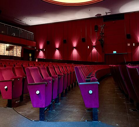 Feste Stuhlreihen, die Sitze mit rotem Samt überzogen. Die hintere Wand des Saales ist ebenfalls rot, 4 Lampen sind im Bildausschnitt zu sehen. Links ein Rang mit silberfarbenem Geländer.