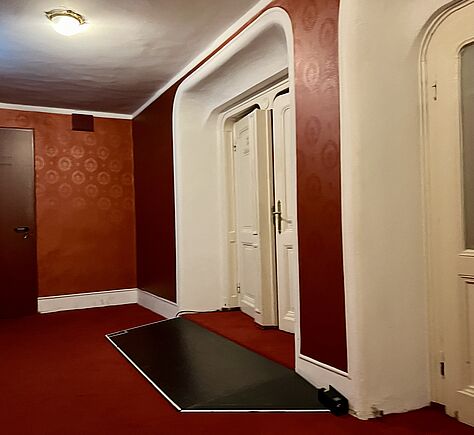 Rechts im Bild, eine geschlossene, weiße Tür mit zwei Flügeln. Vor der Tür gleicht eine Rampe aus dunklen Holz, eine Stufe aus. Auf dem Boden liegt roter Teppichboden, auch die Wände sind rot tapeziert.