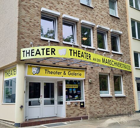 In einem mehrstöckigen Gebäude mit heller Backsteinfassade befindet sich der Eingang. An der Hausecke ist eine Säule mit einer Klingel. Über dem Eingang ist ein gelbes, geschwungenes Vordach mit der Aufschrift 'THEATER THEATER AN DER MARSCHNERSTRASSE' in schwarzen Großbuchstaben.
