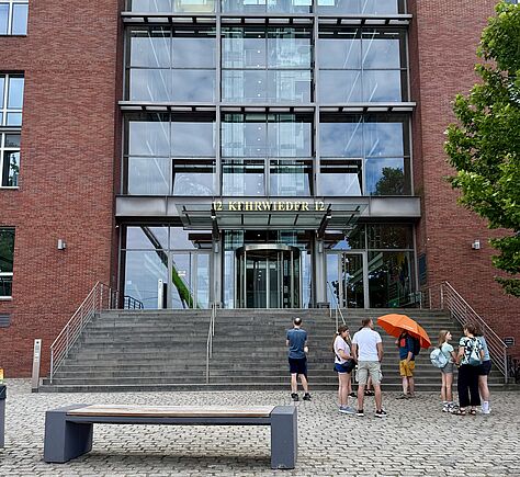 Vor einem modernen Backsteingebäude mit großer Glasfassade stehen mehrere Personen auf einem gepflasterten Platz. Eine Person trägt einen großen orangefarbenen Schirm. Über dem Eingang ist in gelben Buchstaben die Adresse '12 KEHRWIEDER 12' angebracht. Zum Eingang führt eine sehr breite Treppe mit 15 Stufen mit Handläufen rechts, links und zwei weiteren. Eine Rufsäule befindet sich links am Fuß der Treppe. Ein Baum mit grünem Laub ragt am rechten Rand ins Bild. Im Vordergrund steht eine schlichte Bank aus Holz und Metall.