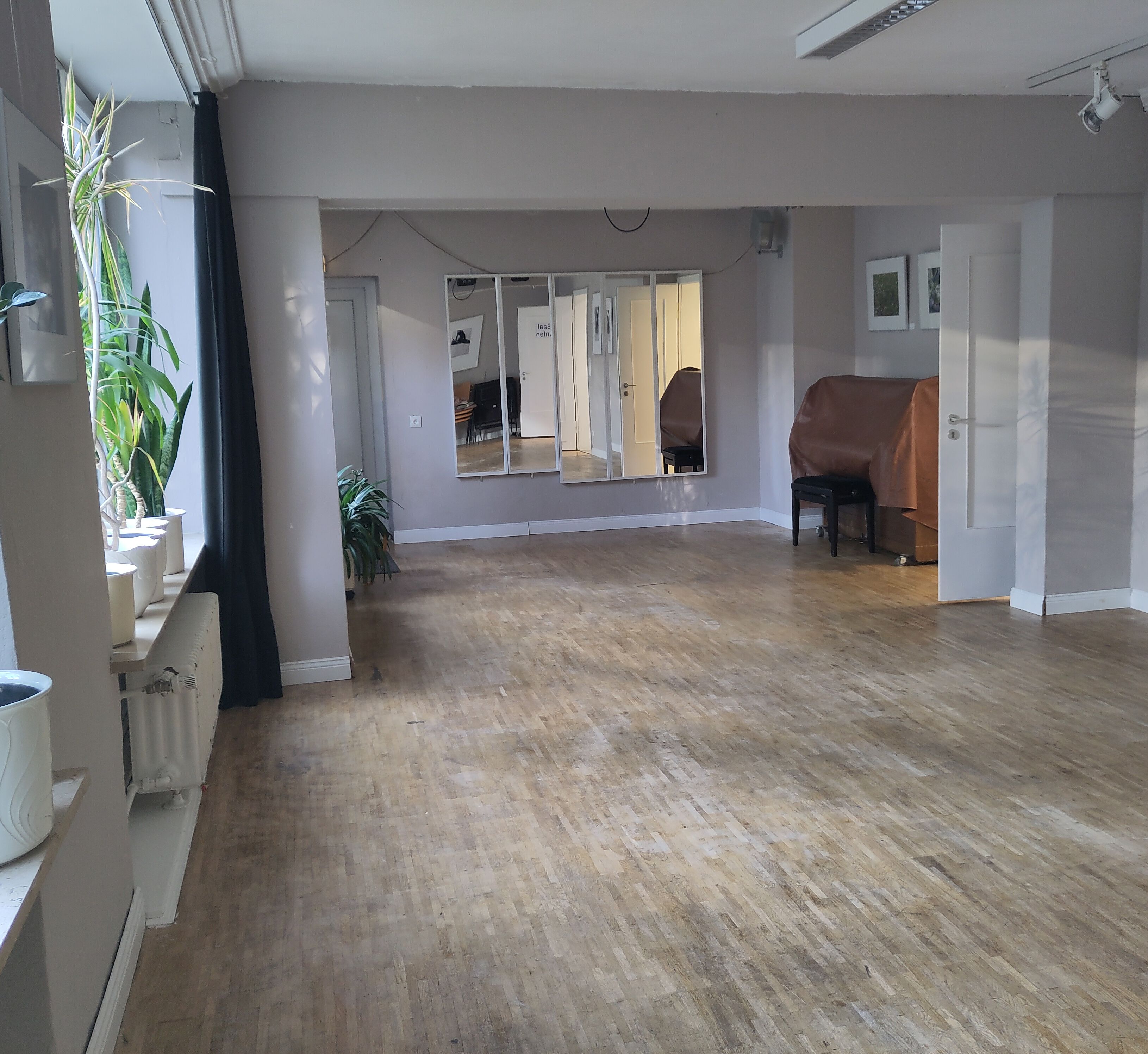 Ein 2-teilger großer sonnendurchfluteter Raum. An der hinteren Wand hängen 2 unterschiedlich große Spiegel, in der rechten Ecke steht ein abgedecktes Klavier. Links auf den Fensterbänken große Pflanzen. Der Boden ist ein heller Parkettbelag, die Wände sind weiß, rechts hängen Bilderrahmen in beiden Teilen des Raumes. Sie sind durch einen breiten Durchbruch getrennt.