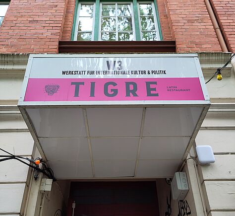 Eingang mit roter Tür unter einem Schild mit der Aufschrift 'V3 WERKSTATT FÜR INTERNATIONALE KULTUR & POLITIK' und 'TIGRE LATIN RESTAURANT'