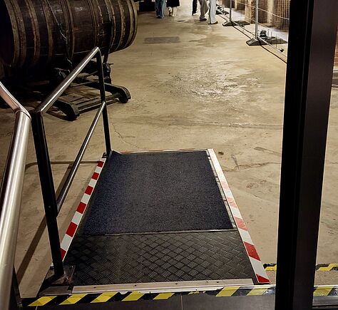 Eine kurze Rampe mit dunkler, rutschfester Oberfläche und Metallrand. An den Kanten sind rot-weiße sowie gelb-schwarze Markierungen angebracht, seitlich verläuft ein Metallgeländer.