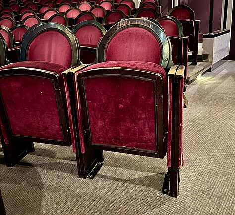 Im Vordergrund sind zwei rote Theaterstühle zu sehen. Die Sitzflächen sind nach oben geklappt im Hintergrund die anderen Stuhlreihen