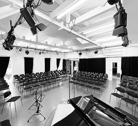 Schwarz-weiß-Foto des Saales. Blöcke mit Stuhlreihen stehen im Halbkreis. im Vordergrund ein Flügel, daneben ein Notenpult und ein Mikrofonständer. Die Decke ist mit mehreren Scheinwerfern und Traversen ausgerüstet, an den Wänden gibt es Fenster mit dunklen Vorhängen. Im Hintergrund ist ein Mensch am Rand des Saals zu sehen. 