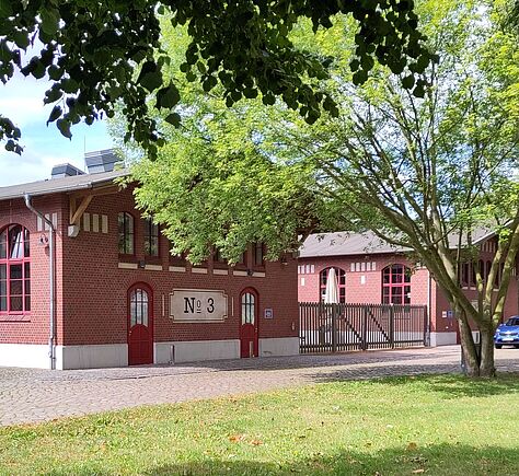 Im Vordergrund erstreckt sich eine Rasenfläche, die von einem Baum mit dichten grünen Blättern teilweise beschattet wird. Dahinter stehen zwei Backsteingebäude mit großen, Rundbogenfenstern. An dem linken Gebäude ist zwischen zwei roten Türen ein rechteckiges helles Schild mit der Aufschrift 'No. 3' angebracht. Ein Metallzaun befindet sich zwischen den beiden Gebäuden. Davor ist ein breiter gepflasterter Weg, am Rand rechts steht ein Baum.