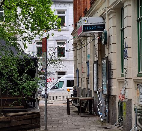 Tische und Stühle am rechten Rand vor einer Hauswand. Daran hängt ein rechteckiges Schild mit der Aufschrift 'TIGRE' in Großbuchstaben. Im Hintergrund steht ein Transporter vor einem weiteren Wohnhaus. Über dem Durchgang sind Lichterketten gespannt, links Bäume mit grünem Laub.