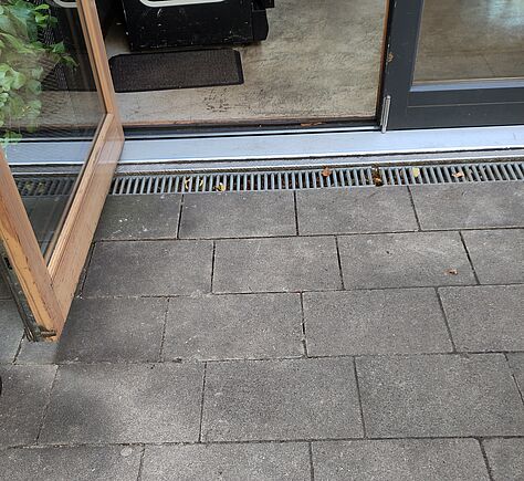 Steinplatten einer Terrasse. Die linke Seite der Doppeltür ist nach außen geöffnet. Im Innenraum liegt eine Fußmatte. Vor der Tür verläuft eine Regenablaufrinne aus Metall.