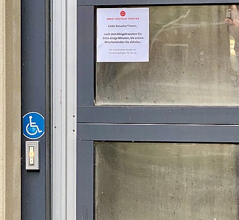 Nahaufnahme von der Klingel. Darüber ein blaues Rollstuhlsymbol. Auf der Tür ein Schild mit dem Text, dass es einige Minuten dauert, bis eine Mitarbeiterin nach dem klingeln kommt.
