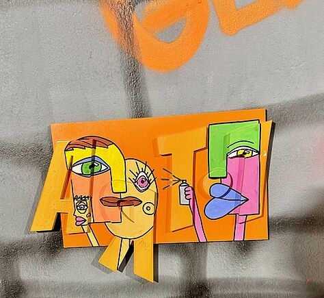 Ein oranges Schild an einer Wand, die besprüht ist. Auf dem Schild steht ART, dabei sind beim Buchstaben A zwei gezeichnete Köpfe und ei T ein seitliches Profil einer Figur im Comicstil