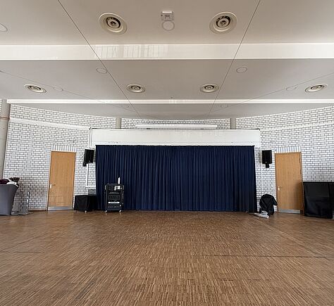 Ein großer Raum mit Holzboden und einer Bühne mit dunkelblauem Vorhang in der Mitte. Links und rechts neben der Bühne sind Türen in der weißen Backsteinwand. Der Raum wirkt offen und ist für Veranstaltungen geeignet.